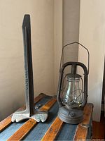 Iron industrial stand next to vintage oil lantern on wooden surface, showing full view of stand's height and lantern shape.