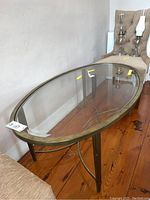 Oval glass top coffee table with brass colored metal base resting on wooden floor, beside beige upholstered chairs and lamps in background.