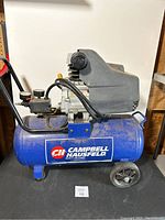 Front side view of the air compressor showing Campbell Hausfeld blue tank with logo, motor on top, black protective cover, and wheels.