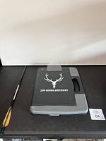 Archery case with antler skull logo, closed, alongside an arrow and lighter on black surface