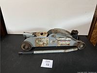 Angle showing side and top of gray metal car hydraulic jack with handle lying beside it