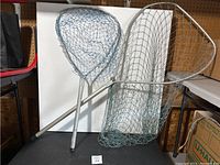 Two fishing nets leaning against a white board on a table, showing the netting styles and metal handles.