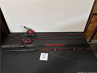 Two fishing rods placed side by side horizontally showing red reels and black handles on a dark surface.