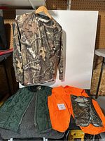 Hunting clothing lot consisting of three items displayed on hangers and laid out: camouflage shirt, orange vest, quilted green vest.