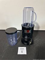 Magic Bullet Single Shot+ blender with base, large blending cup with handle and lid, and smaller cup with lid on black surface.