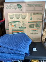 Photo of folded blue quilted moving blankets stacked with U-Haul branded cardboard moving boxes in background