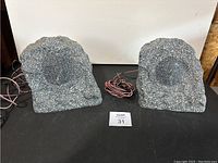 Pair of gray rock-like outdoor speakers on a dark surface with wire between them.
