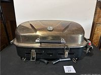 Front view of the portable BBQ grill showing metal lid, locking latches, and propane gas connector.