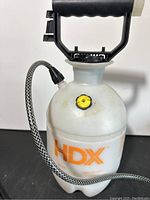 Front view of white HDX branded plastic sprayer tank with black pump handle and braided hose attached.