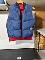 Front view of vintage navy blue Sports Chief insulated vest with red interior lining, hanging from wooden hanger, on pegboard background.