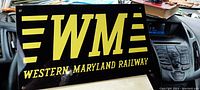 Front view of black and yellow Western Maryland Railway sign on a car dashboard showing the entire design with 'WM' logo and text.