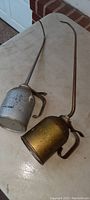 Two large antique metal machinery oiler cans resting on a white surface, showing their metal spouts, thumb pumps, and handles. One is silver colored, the other brass/golden tinted.