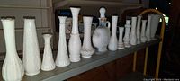 Side view of 16 white milk glass vases and one decanter bottle placed in a row on a shelf, showing varying height and raised patterns.