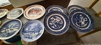 Photo showing antique plates with blue and white scenic designs, including winter and traditional village scenes, some with intricate border patterns.