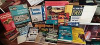 Photo showing a box full of colorful vintage automotive catalogs and brochures from brands including Chevrolet, Rod & Custom, Midas, and others, featuring mid-century car illustrations and product listings.