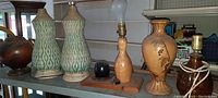 Photo showing five lamp bases of various mid century art deco designs on shelf; includes green patterned ceramic lamps, brown ceramic decorative vase-style lamp base, wood lamp base, and dark brown lamp base with electrical cord; no shades seen