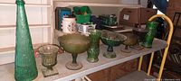 Nine antique green art glass pieces arranged on a shelf showing variety in shapes and textures including tall vases and bowls.