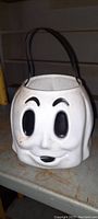 Front view of white ghost-shaped hard plastic trick or treat bucket with black painted face and handle