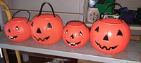Four orange plastic pumpkin-head trick or treat buckets on a white surface with different carved face designs and black handles