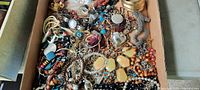 Photo showing a flat box filled with assorted costume jewelry including multiple necklaces and bracelets in various colors and materials.
