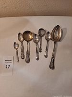 Seven sterling silver spoons and ladle laid out on white cloth, showing front patterns.
