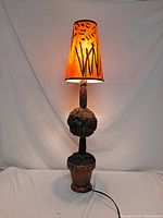 Full view of the illuminated Arts and Craft style lamp showing the pot base, faux greenery, tree trunk and decorated shade.