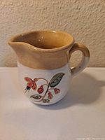 Photo showing front side of small ceramic creamer pitcher with hand-painted floral design and tan upper rim.