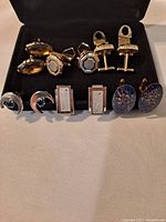 Lot of 6 pairs of vintage cuff links arranged on black display tray showing a variety of silver and gold tone styles including geometric, textured, crescent moon, and glittery designs.