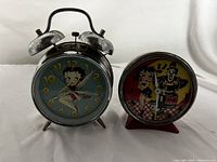 Front view of two vintage Betty Boop alarm clocks placed side by side, showing full faces and wear on metal and paint surfaces.