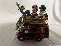 Front and side view of the red musical bus figurine featuring Goofy, Minnie Mouse, Mickey Mouse, Donald Duck, and Pluto with the 'SEE THE STARS HOMES!' banner on the side.