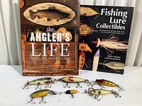 Photo of six wooden fishing lures of various sizes arranged in front of two hardcover books about fishing and lure collecting.