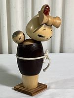 Front view of the Kokeshi doll showing the mother figure with facial details and baby on the back, with a white cord tied around the body and a wooden base