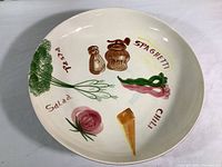 Overhead view of large white ceramic serving bowl with painted vegetables, salt and pepper shakers, and various food type labels inside.