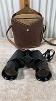 Binoculars in front of open brown leather carrying case with strap, showing overall item condition and size