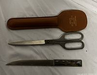 Photo showing brown leather sheath, metal scissors with black handles, and metal letter opener.