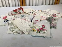Overview of a pile of over two dozen vintage linen handkerchiefs mostly white or off-white with colorful floral embroidery and printed patterns.