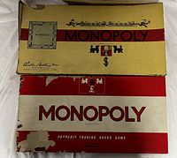 Photo showing two vintage Monopoly board game boxes stacked, with visible damage and wear on boxes.