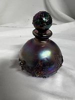 Front view of the perfume bottle showing spherical shape, iridescent finish, and lava texture on glass with round stopper.