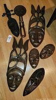 Seven African wood masks arranged with one carved table leg, all on a wooden floor showing detailed carving and finishes.