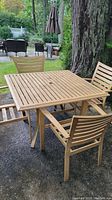 Square aluminum outdoor dining table and four matching chairs on gravel near large tree showing weathered tan/beige finish