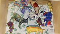 Top down view showing eleven colorful ceramic cow figurines laid out on a floral patterned cloth, revealing diverse artistic styles and colors.