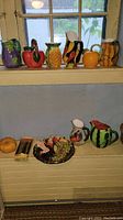 Wide shot of all ceramic fruit pitchers and decorative items arranged on window sill and shelf showing variety of fruit shapes including pineapple, pumpkin, pepper, watermelon, and grape clusters