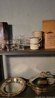 Photo showing Dansk Nielstoft boxed glassware, clear glass dessert bowls, ceramic cups, and silver plated serving dish and platter on metal shelf.