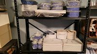 Shelf with various dishes, cups, saucers, teapot, and serving pieces mostly wrapped or in protective packaging.