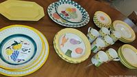 Full view of ceramic dishes and mugs featuring various fruit designs and yellow accents on wooden table.