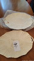 Two Lenox ceramic platters on a wooden table; one cream colored with decorative cut-out handles and floral embossing, the other off-white with solid handles and embossed design, both with glossy finish, bubble wrap nearby.