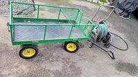 Green metal rolling cart with mesh sides and yellow wheels shown on pavement, accompanied by garden hose reel with hose and spray nozzle