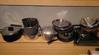Photo showing a collection of various pots, pans, strainers, and cooking utensils lined up on a ledge against a wall.
