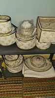 Stacked fabric bags with clear tops containing various Rose China porcelain pieces, some wrapped in plastic, on upper and lower shelves.