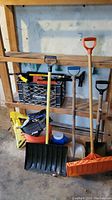 Three snow shovels and a snow pusher lean against a wooden shelf with a black crate containing small snow removal tools, a yellow bag of snow salt, gloves, and an orange bucket on the floor.